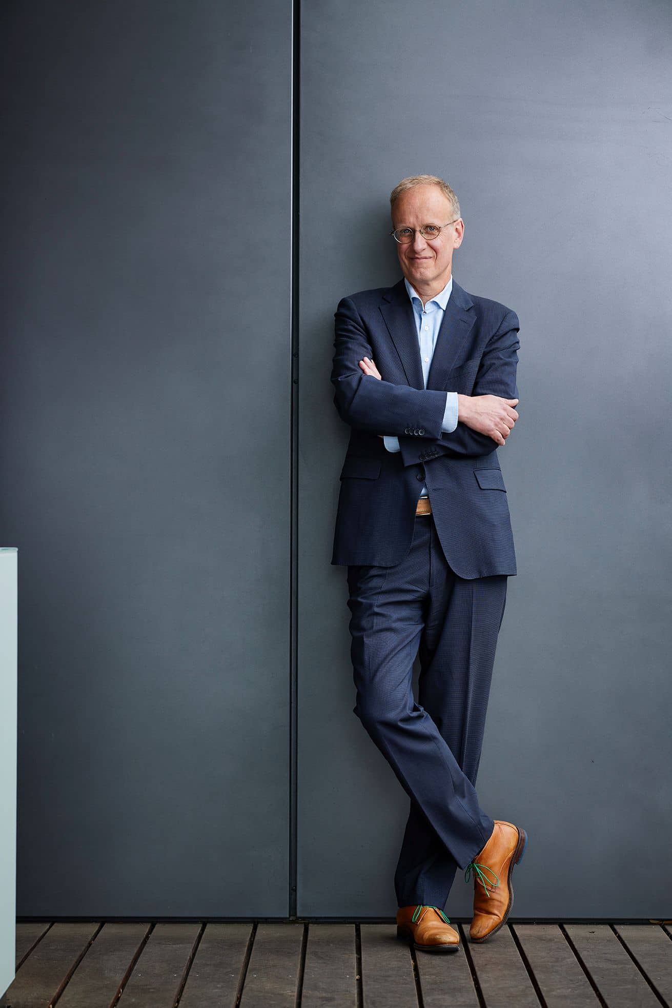 Een man met wat grijzig haar, een bril en in pak, hij leunt tegen een muur