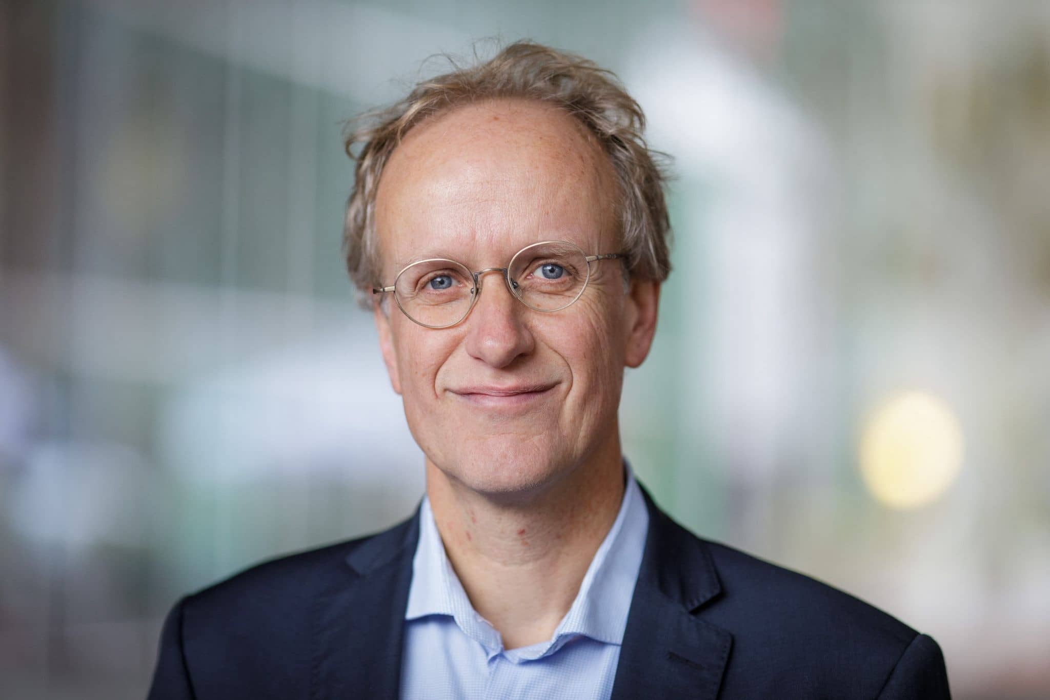 Portret vanaf de schouders, een man met wat grijzig haar en bril, de achtergrond is wazig